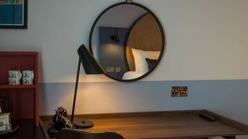 A room with a wooden desk, a round mirror, a red shelf with mugs, and a lamp.