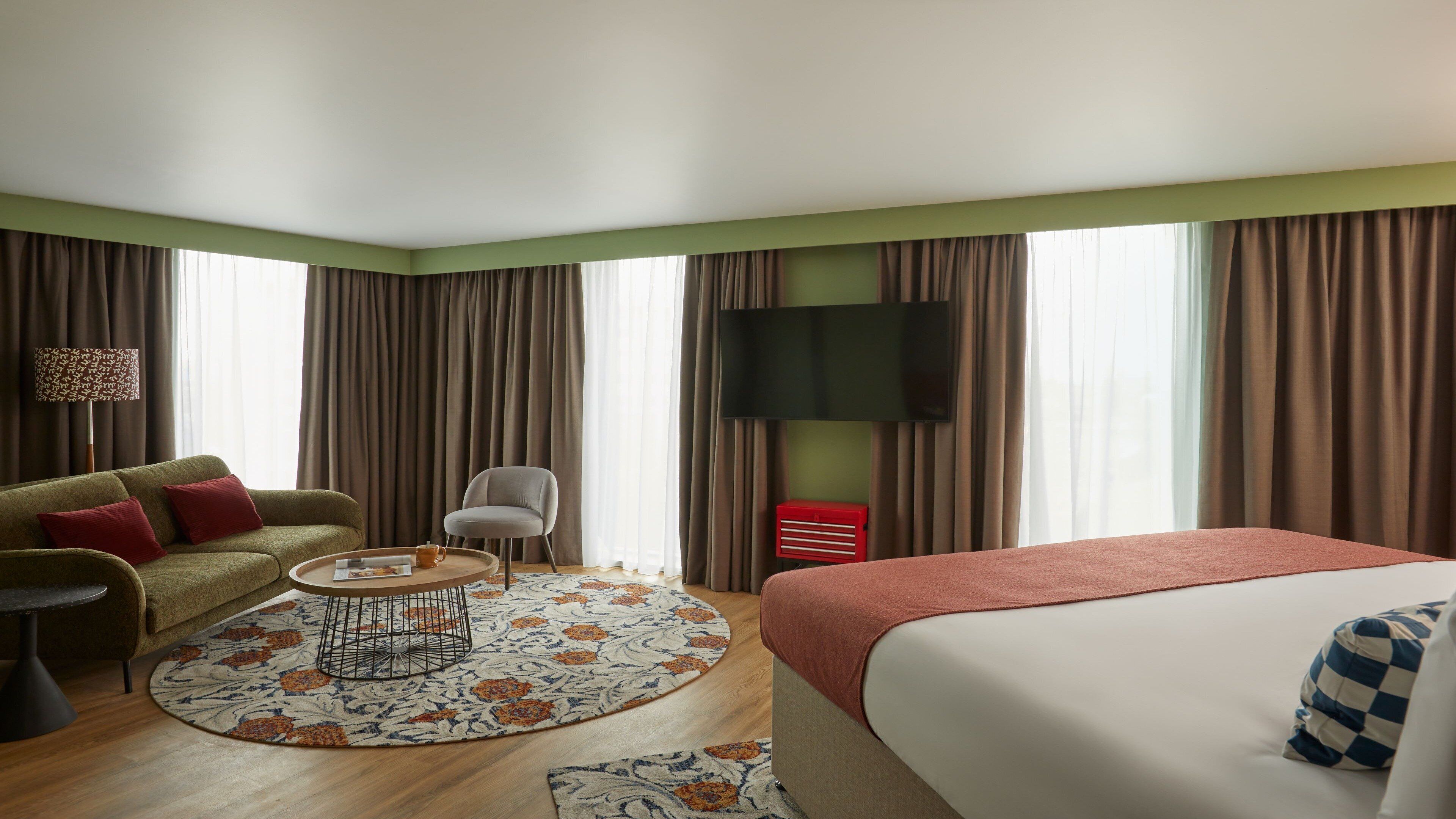 A hotel room with a bed, a sofa, a chair, a coffee table, a TV, and curtains.
