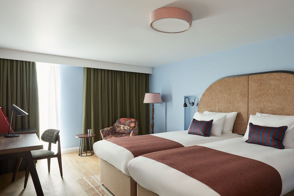 A hotel room with a large bed, a desk with a lamp, a chair, and a window with curtains.