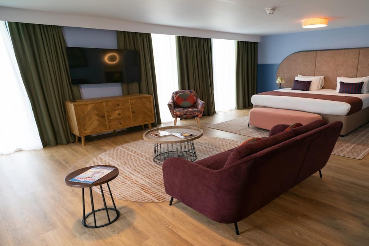 A modern hotel room with a large bed, a sofa, a small table, and a TV mounted on the wall.