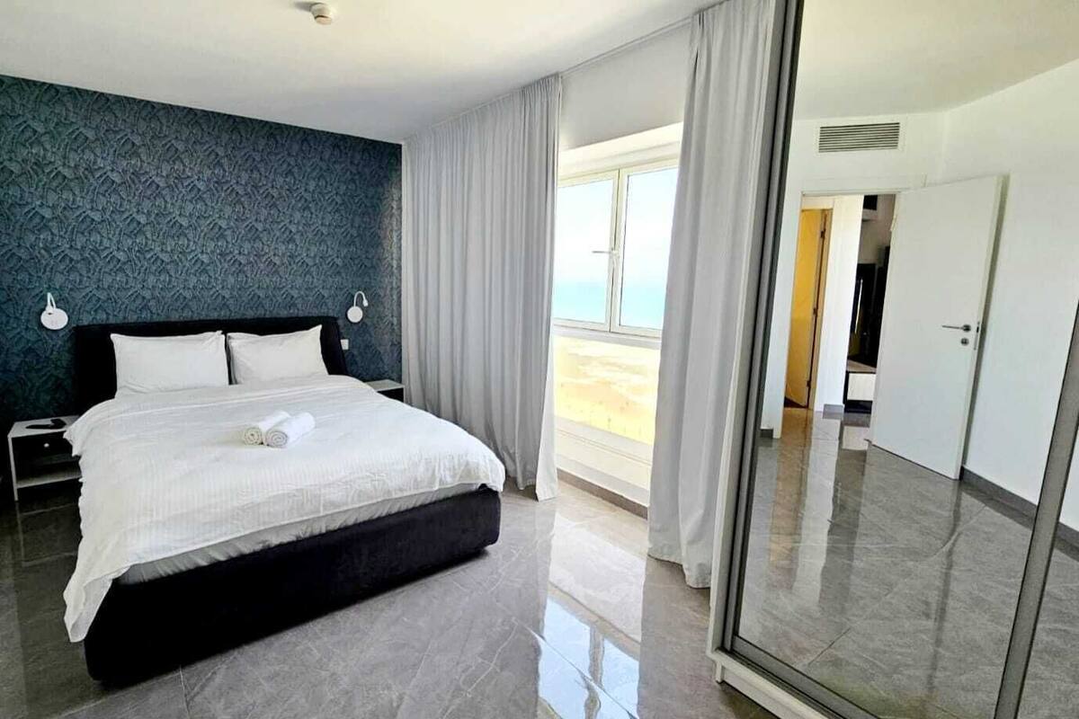A modern hotel room with a large bed, a view of the beach, and a sliding glass door leading to a balcony.