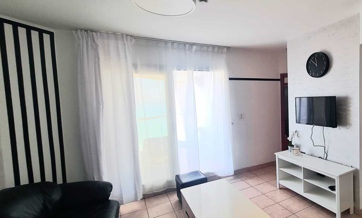 Une pièce comprenant un fauteuil en cuir noir, une table blanche, un téléviseur fixé au mur et une fenêtre ornée de rideaux légers.