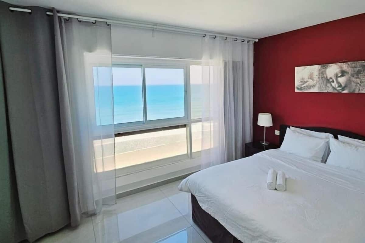 A hotel room with a bed, a nightstand, a lamp, a painting on the wall, and a view of the ocean through the window.