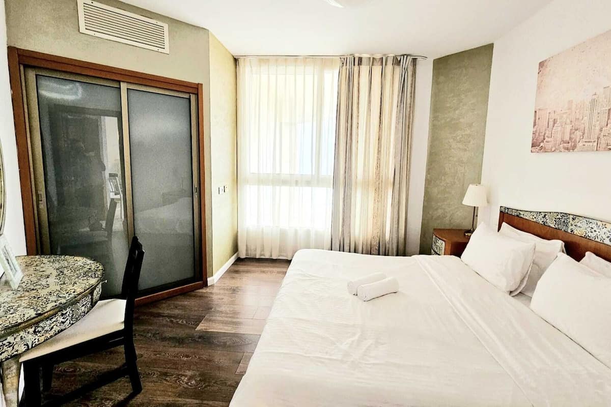 A hotel room with a bed, a desk, a chair, a window with curtains, and a painting on the wall.
