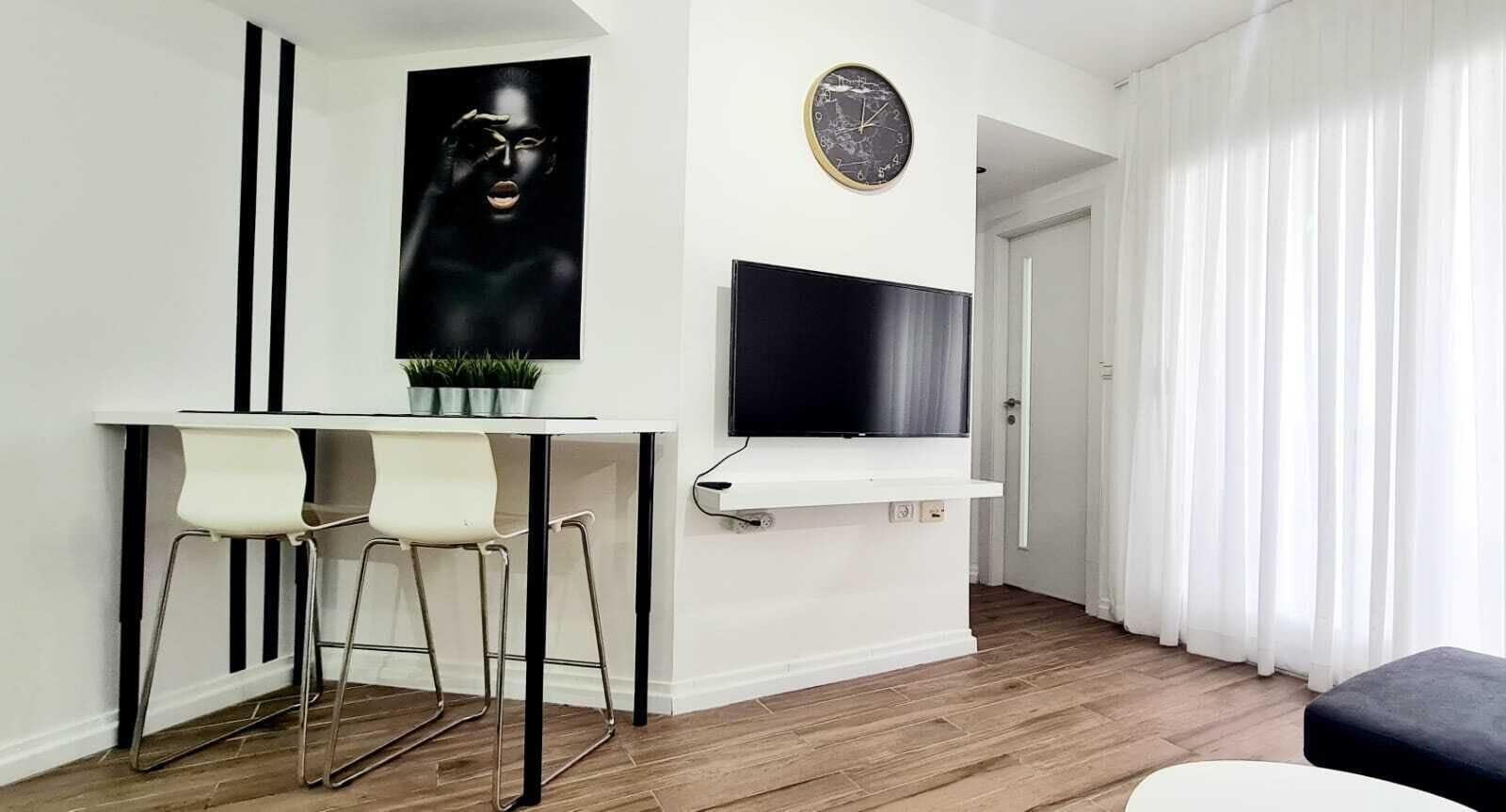 Un salon moderne avec un mur blanc, un téléviseur à écran plat fixé au-dessus d’une console blanche, deux tabourets de bar blancs, une plante en pot et une grande fenêtre ornée de rideaux blancs.
