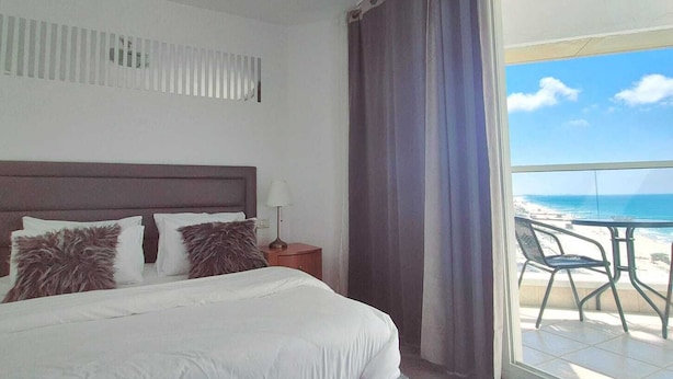 A hotel room with a bed, a nightstand, a lamp, a window with curtains, and a view of the beach. - SHaYaSH apartments Leonardo Bat-Yam (Bat Yam)