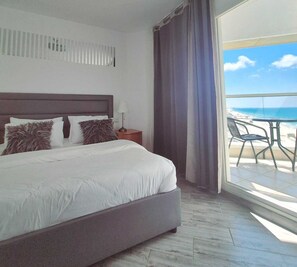 Habitación de hotel con cama, mesita de noche, lámpara, ventana con cortinas y vista a la playa.