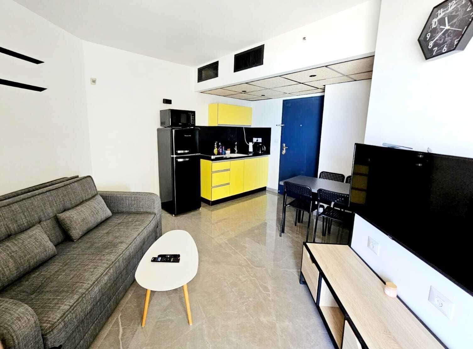 A compact living space with a grey sofa, a small white table, a black TV, a kitchen area with yellow cabinets, and a blue door.
