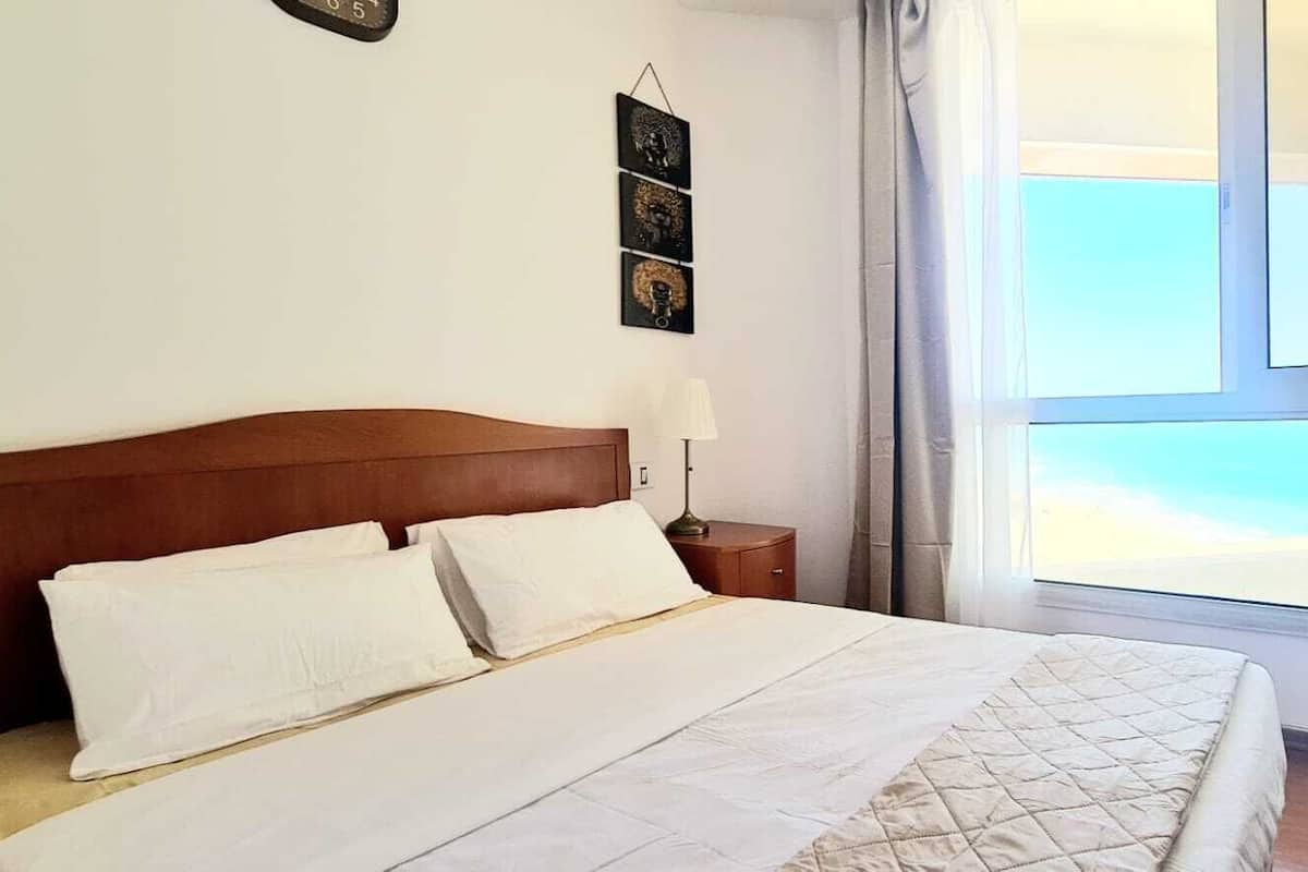 A neatly made bed with white linens in a room with a wooden headboard, a bedside table with a lamp, and a view of the sky through a window.
