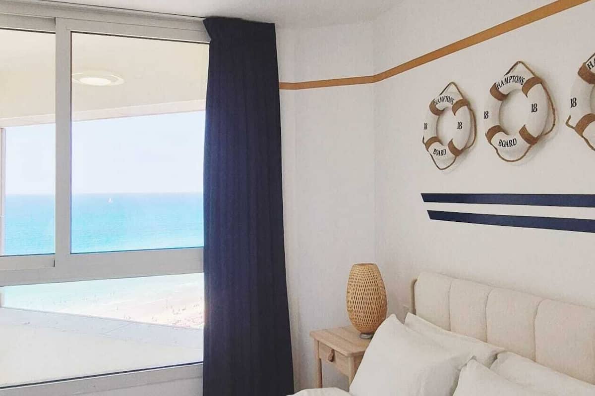A hotel room with a large bed, a nightstand, a decorative wall piece, and a view of the beach.