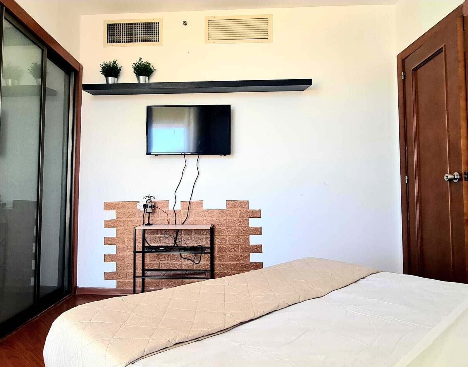 Une chambre à coucher avec un lit, un téléviseur fixé au mur, une étagère avec des plantes et une porte.