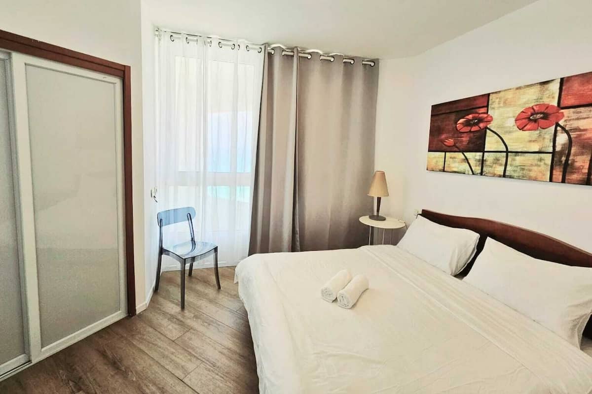 A hotel room with a bed, a chair, a nightstand with a lamp, and a painting on the wall.