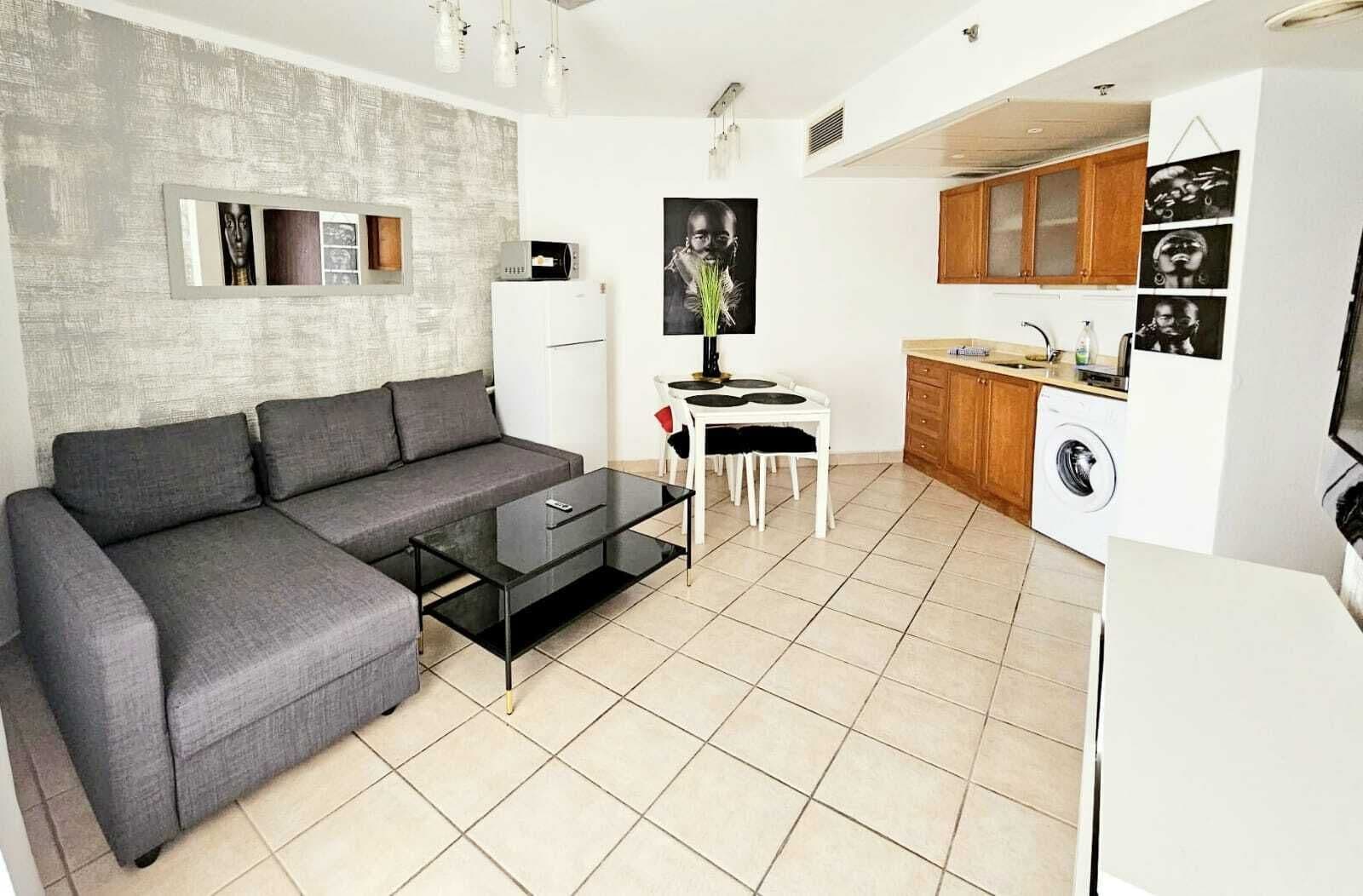 A modern living room with a grey sofa, a glass coffee table, and a dining table with chairs. A kitchen with wooden cabinets and a washing machine is visible in the background.