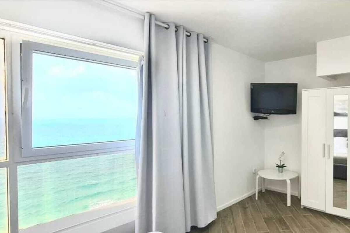 A room with a large window overlooking the sea, a flat-screen TV mounted on the wall, and a small table with a potted plant.