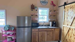 Una cocina con armarios de madera, un refrigerador de acero inoxidable, un estante rosa con utensilios de cocina, un reloj y una ventana con vistas a la vegetación.