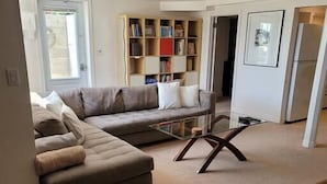 Ein Wohnzimmer mit Ecksofa, Hocker und Glastisch. Eine Bücherwand mit Büchern und einem gerahmten Bild an der Wand.