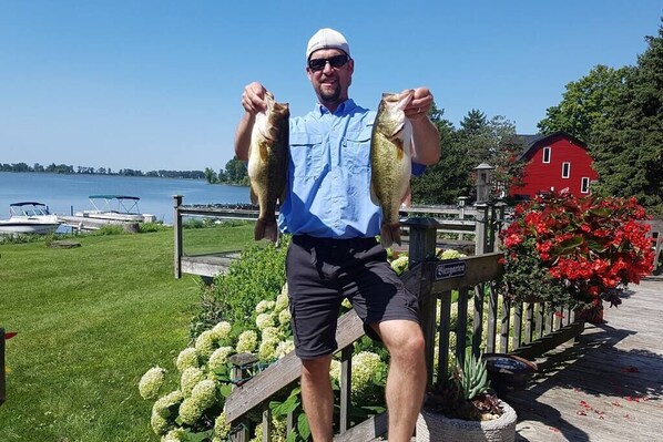 A person holding two fish on a dock with a red barn and a grassy area in the background.