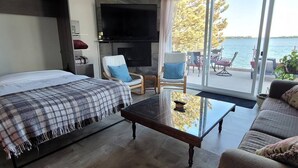 Ein modernes Schlafzimmer mit einem großen Bett, einer Couch, einem Couchtisch und einem Fernseher. Ein Balkon bietet Blick auf das Wasser.