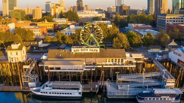 A waterfront cityscape with a prominent Ferris wheel, a marina with boats, and the Rio City Cafe building.