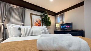 A neatly made bed with a white blanket, a TV on a stand, and a plant in the corner.