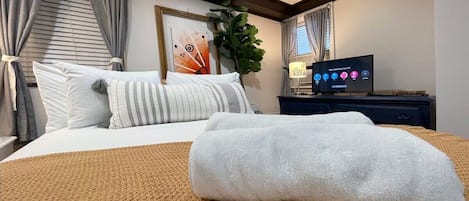 A neatly made bed with a white blanket, a TV on a stand, and a plant in the corner.