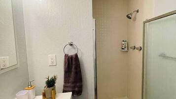 Une salle de bain avec un lavabo blanc, un miroir, un porte-serviettes, une douche avec une porte en verre et une petite plante en pot.