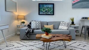 Un salon moderne avec un canapé gris, une table basse en bois, un tapis à motifs et un tableau encadré accroché au mur.