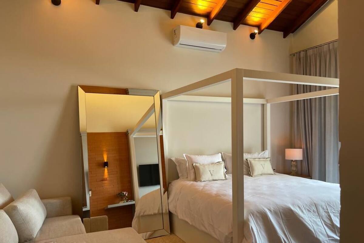 A modern hotel room with a four-poster bed, a sitting area with a sofa, a mirror, and a bedside table.