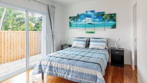 Ein Schlafzimmer mit einem großen Bett, einem Deckenventilator, einer Schiebetür zu einem Balkon und einem Wandbild mit einer Strandansicht.