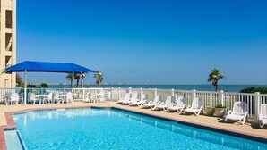A clear blue pool with white lounge chairs and umbrellas, surrounded by a white fence, overlooking a beach.