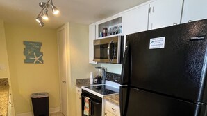 A compact kitchen with white cabinets, a black refrigerator, a microwave, and an oven. There is a trash can and a small rug on the floor.