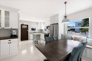A modern kitchen with white cabinetry, a large wooden dining table, and a view of the outdoors through a window.