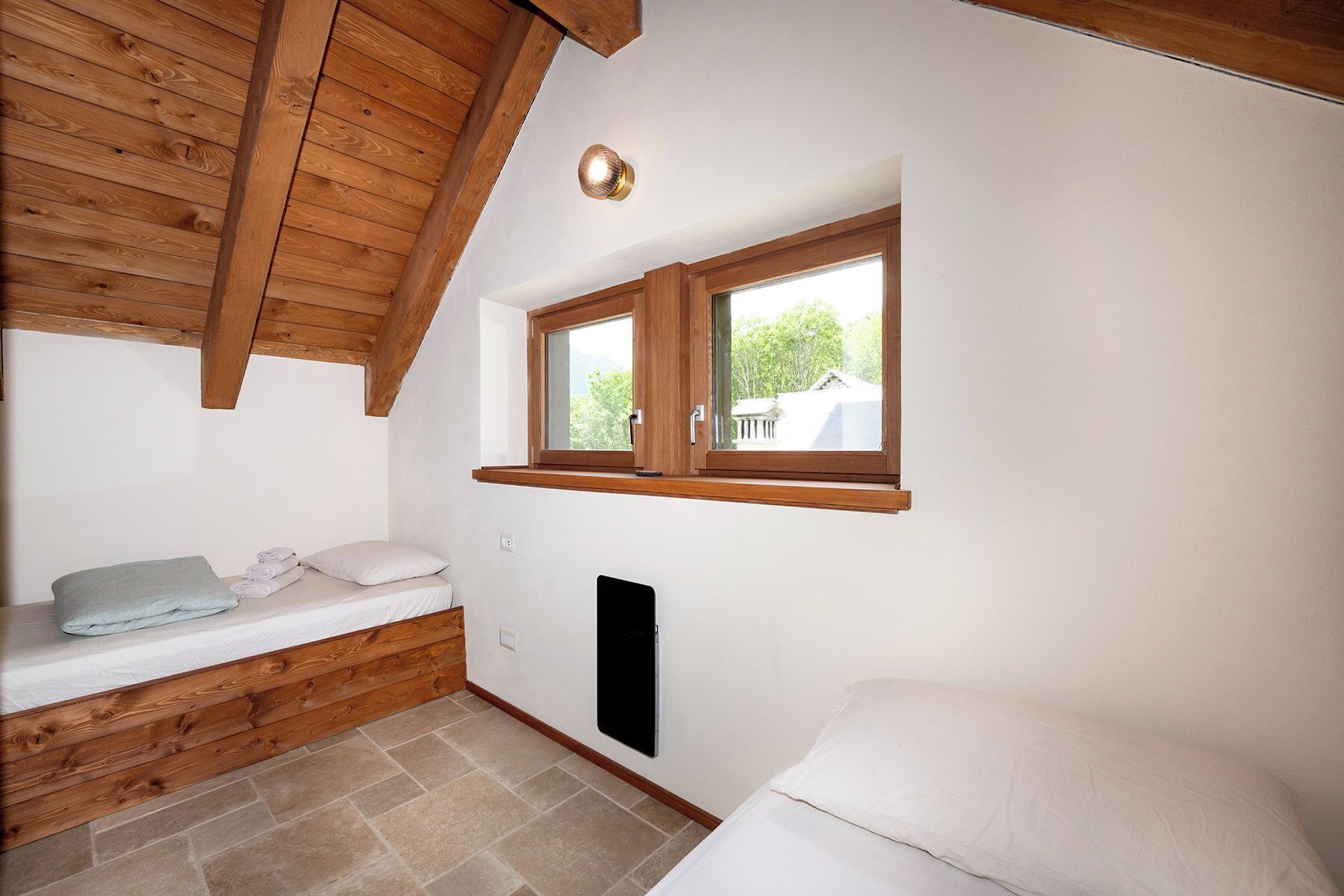 Une petite pièce accueillante avec un plafond en pente, une fenêtre avec des cadres en bois, un banc intégré doté de coussins et un luminaire fixé au mur.