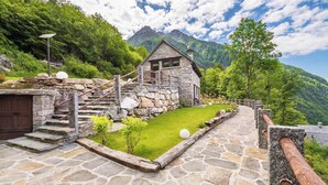 A stone house with a stone staircase leading to a wooden deck, surrounded by lush greenery and mountains in the background. - Baita Athili - Crodini Village (Piemonte)