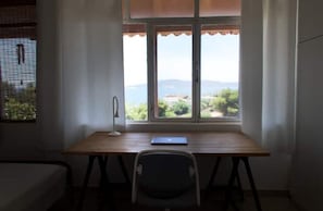 Une pièce avec un bureau en bois, une chaise et un ordinateur portable. Il y a une fenêtre donnant sur un paysage verdoyant et vallonné.
