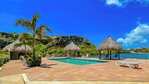 A pool area with lounge chairs and thatched umbrellas, surrounded by greenery and a view of the sea.