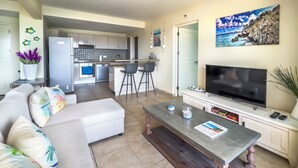 A modern living room with a sofa, a coffee table, and a TV. There is a kitchen in the background with a fridge, stove, and bar stools.