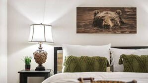 A bedroom with a bed, a bedside table with a lamp, a wall-mounted picture of a bear, and a woven basket on the bed. - Center Hill Serenity (Silver Point)