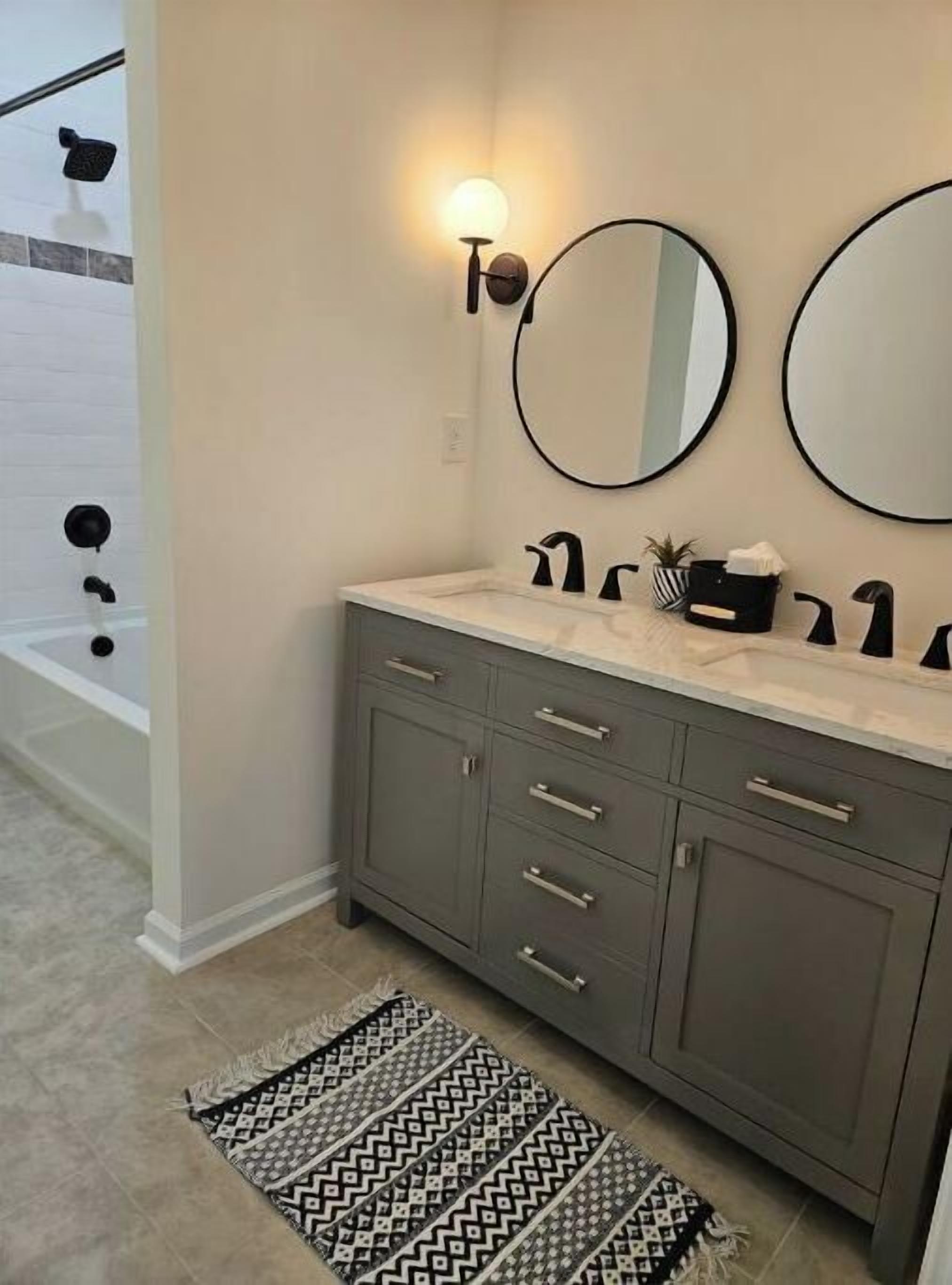 A bathroom with a vanity, two round mirrors, a wall-mounted light, and a patterned rug.