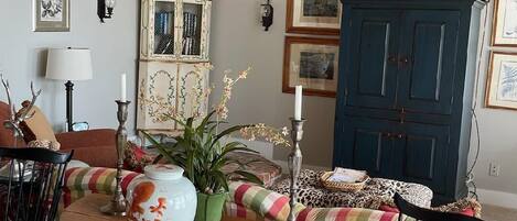 Een gezellige woonkamer met een houten plafond, een geruite bank, een ronde tafel met een vaas en kaars, en een blauwe kast met ingelijste foto's.