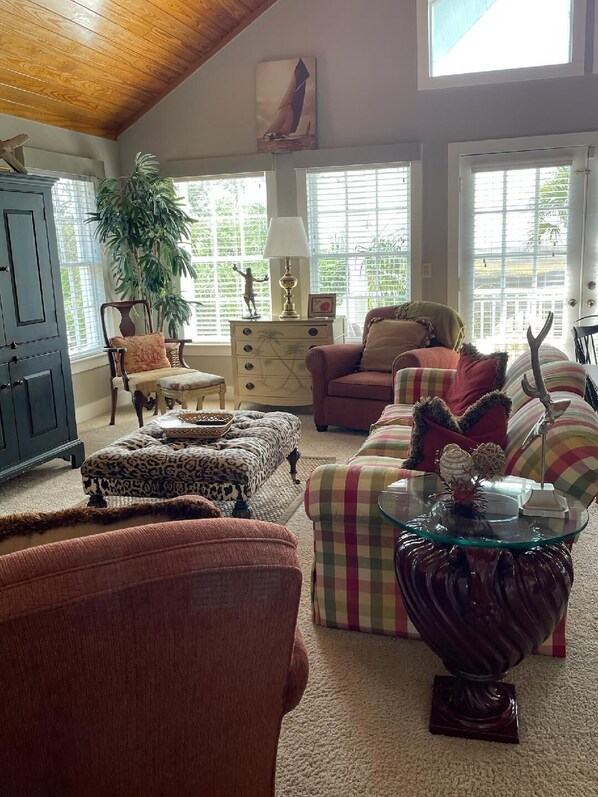 A cozy living room with a wooden ceiling, a large window with blinds, a sofa, a chair, and a decorative glass table.
