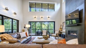 A spacious living room with a large leather sofa, a coffee table, and a fireplace. There are multiple windows providing natural light and views of the outdoors.