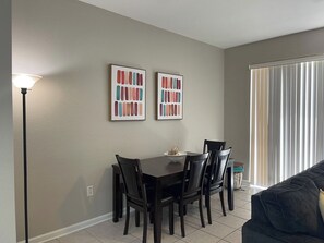 A dining area with a table and chairs, two framed color swatches on the wall, a floor lamp, and a window with blinds. - Disney Area - Heated Pool - 3 Bedroom Townhome (Kissimmee)