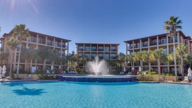 A hotel with a central pool and a fountain, surrounded by palm trees and multiple-story buildings. - Amazing Moonlight Escape- (Panama City Beach)