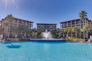 A hotel with a central pool and a fountain, surrounded by palm trees and multiple-story buildings.