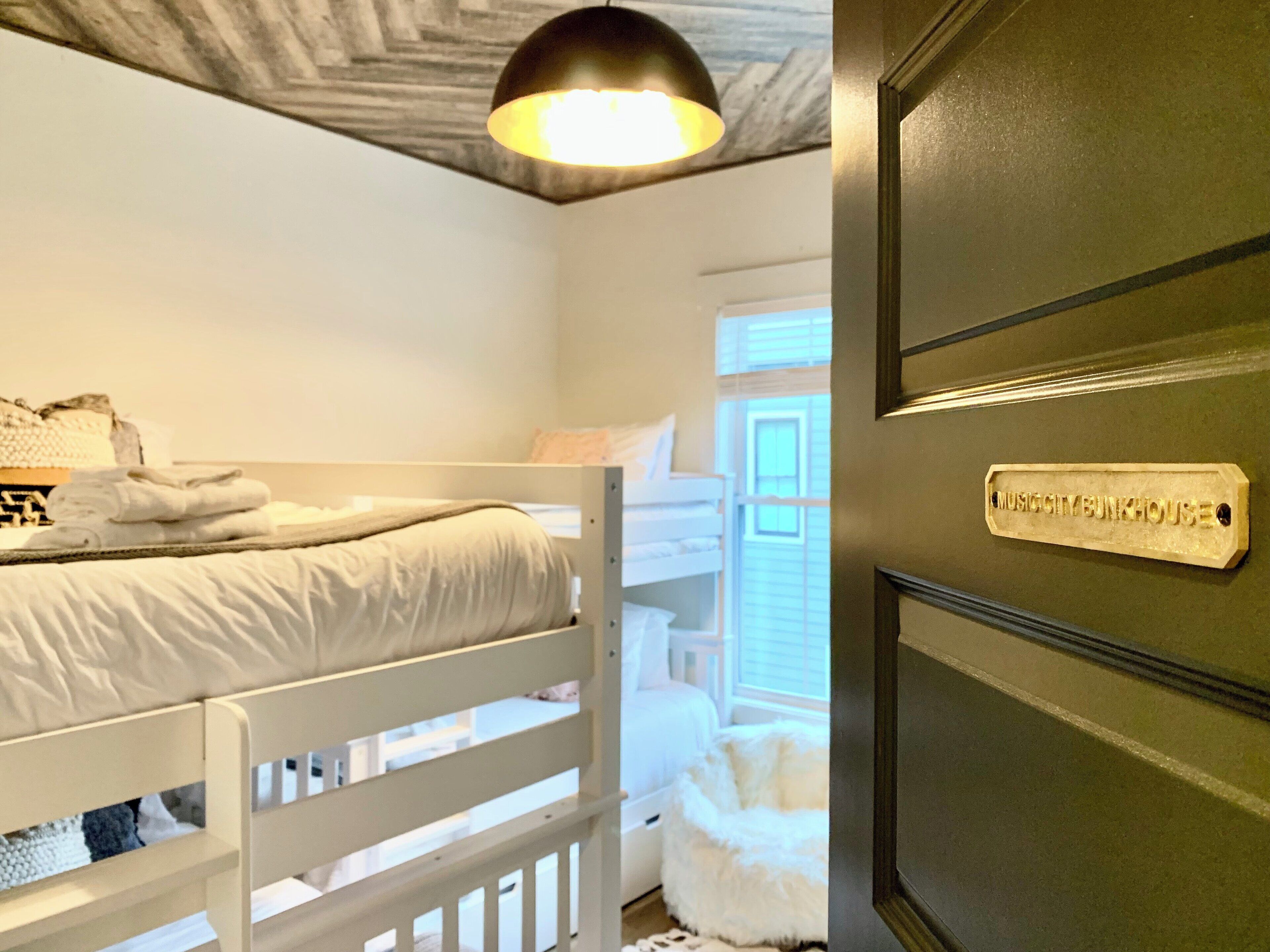 Une chambre avec un lit superposé, une chaise blanche, un pouf blanc moelleux et une porte sur laquelle est affiché un panneau indiquant « MUSIC CITY BUNKHOUSE ».