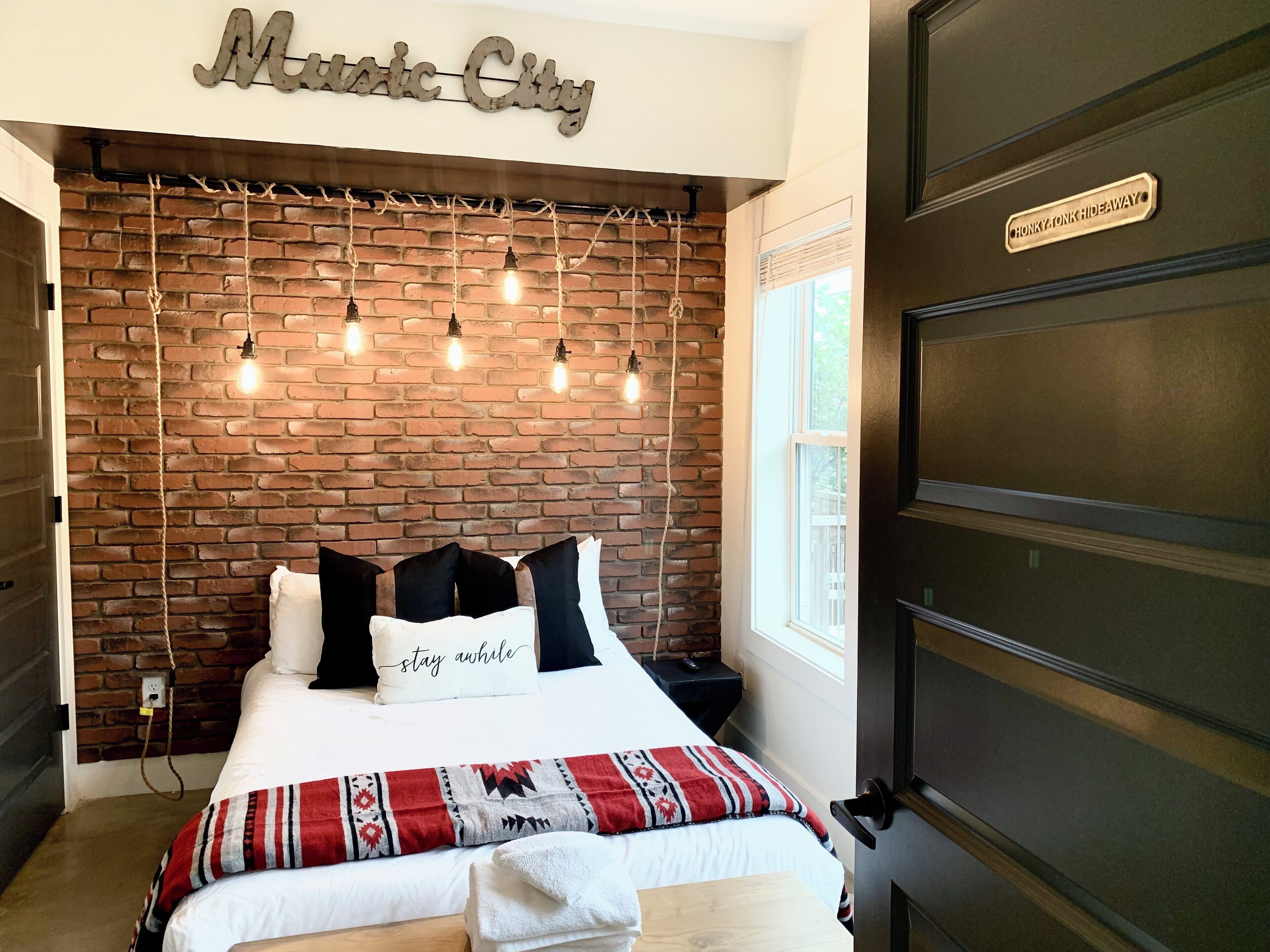 A bedroom with a brick wall, a bed with a patterned blanket, a sign reading "Music City", and a nightstand with a lamp.