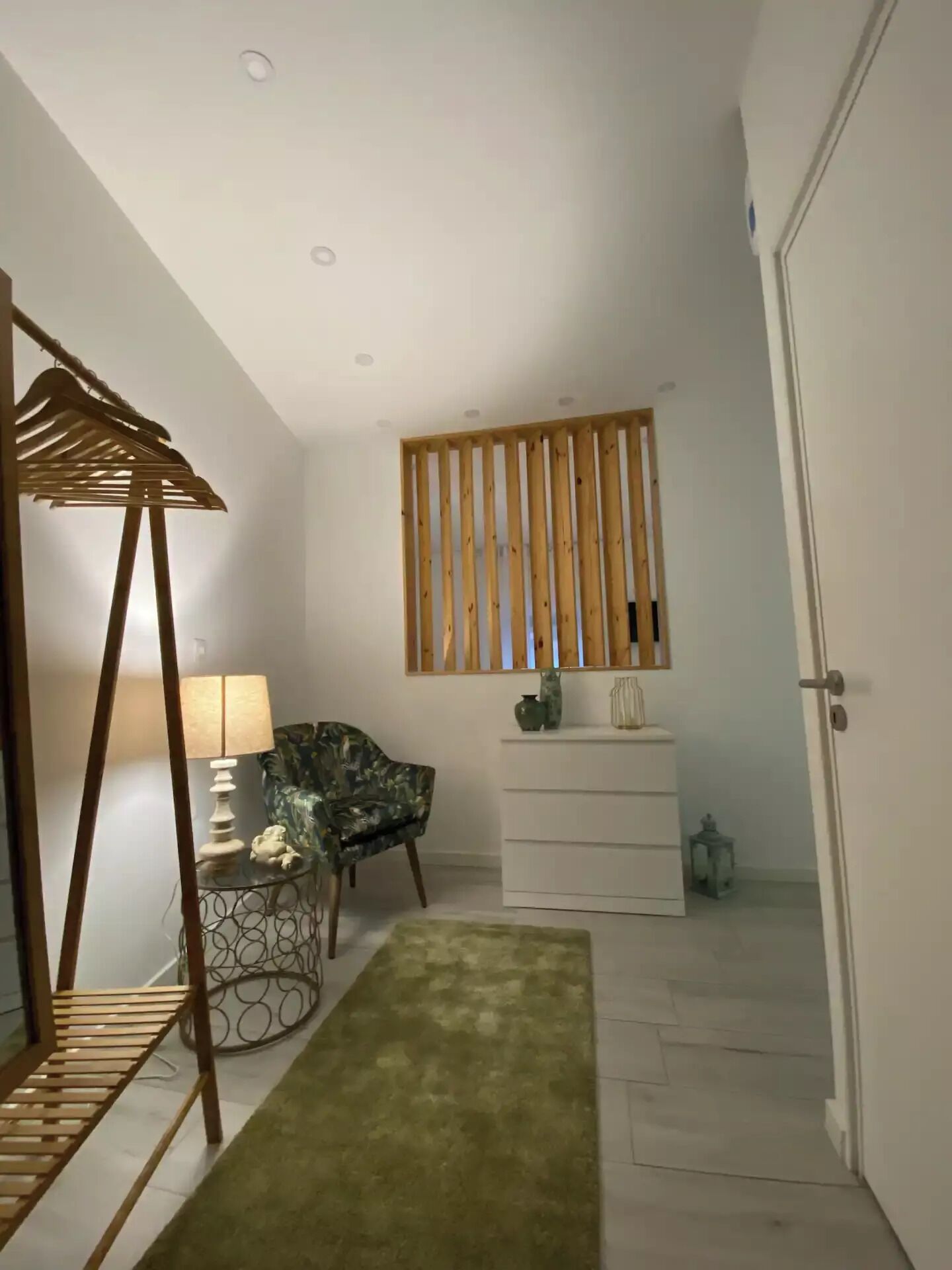 Un couloir avec une cloison faite de lattes de bois, un tapis vert, une chaise, une petite table avec une lampe et une commode blanche.
