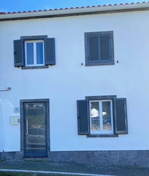 Un edificio de dos pisos con fachada blanca, tres ventanas con contraventanas y una puerta.