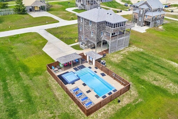 A residential area with a large swimming pool surrounded by a wooden deck, a stone building, and a well-maintained lawn.